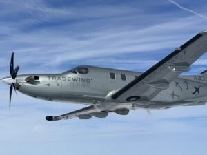 A propeller aircraft labeled 'TRADEWIND' flying against a clear blue sky with wispy clouds.
