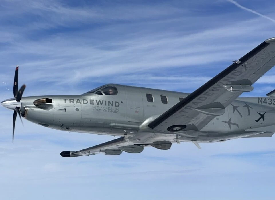 A propeller aircraft labeled 'TRADEWIND' flying against a clear blue sky with wispy clouds.