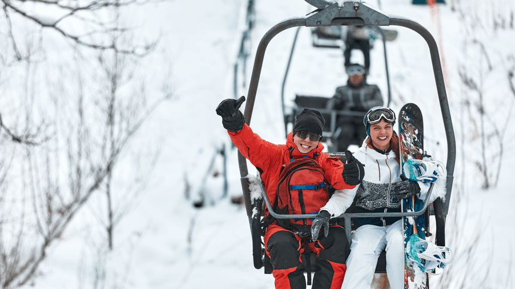 Following a convenient flight to Vermont, two skiers celebrate on a chairlift with snow in the background.