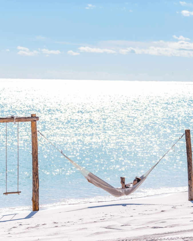 A woman laying in a beach hammock with turquoise waters behind.